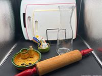 Photo showing five plastic cutting boards in a white stand, clear glass wine decanter, wooden rolling pin with red handles, colorful plastic measuring cups and spoons, ceramic olive bowl with 'Ajo de Puerto Banus' decoration