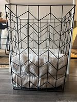 Front view of the black metal bath towel holder with four white towels inside, showing the chevron design of the metal basket.
