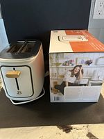 White two-slice toaster with gold lever and B emblem on front next to original box showing product image and branding.