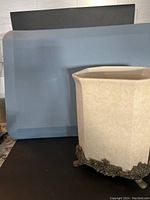 Photo of the blue Kangaroo cushion mat and beige ceramic wastebasket with iron feet and trim against a black background.