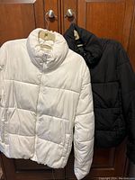 Photo showing both white and black puffer coats hanging side by side on hooks against wooden background.