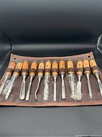 Full set of 12 wooden handle carving tools inside brown foldable pouch, each tool having plastic protective wrap on blade, pouch laid open.
