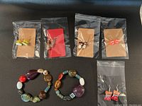 Photo showing two big handmade polished stone bracelets and six pairs of fun novelty earrings in individual clear plastic pouches on a dark surface.