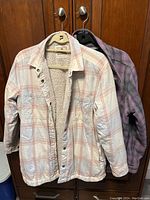 Both jackets hanging on hangers showing front view. Cream plaid jacket is sherpa lined and unbuttoned. Purple plaid jacket is behind cream jacket.