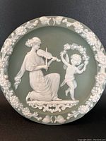 Front view of the Wedgwood Jasperware 5 3/4 inch matte green hanging plate with white classical relief design featuring a woman playing violin and a cherub with flowers.