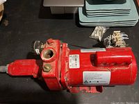 Red electric water pump with attached motor shown from one side on a dark surface with plates in background.