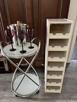 Chromed frame bar cart with glass shelves holding three purple wine glasses and a stainless steel ice bucket with a wine bottle inside. Adjacent to the bar cart is a white vertical wine rack with 14 slots.