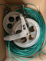 Top down view of the aluminum winding reel and green clothesline wire coiled inside a box showing the handle and circular reel holes.