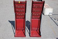 Two red metal car ramps standing upright side by side showing overall shape, bolt holes, and side braces