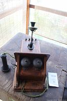 Photo showing top angled view of the antique wooden wall phone, its two metal bells, black mouthpiece, and receiver attached with fabric-wrapped cord.
