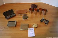 Photo showing all items in the lot displayed on wooden floor: two silver-plated cases, two pairs of vintage eyeglasses, four nested cups with case, opera glasses, celluloid compact, two pens, round metal box.