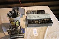 Photo showing plastic canister filled with various drill bits, metal drill bit holder with bits inserted, and large drill bit box set with many drill bits inside