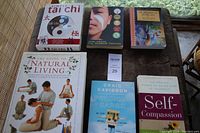 Six self help books on a wooden surface photographed in natural light near a window. Books cover topics including Tai Chi, natural living, emotional health, and self compassion.