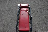 Full view of the mechanics creeper with red vinyl padded surface and black metal frame with wheels underneath, shown outdoors on pavement.
