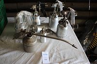 Overview photo showing seven metal spray guns with attached canisters, including a long nozzle oil sprayer and a Campbell Hausfeld sprayer still in plastic wrapping. All placed on a white cloth.