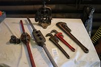 Photo showing the full set of tools laid out on a white surface including pipe wrenches and threading bars with dies