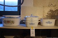 Shelf display of white glass casserole baking dishes with colorful L'Echalote La Marjolaine and blue floral patterns, various sizes, all with lids.