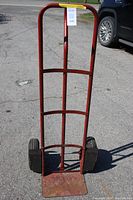Front view of the red steel dolly standing on asphalt pavement, showing overall structure and wheels with welded metal tabs.