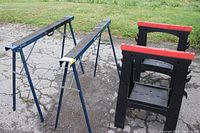 Photo showing two steel sawhorses painted dark blue with folding legs and two black plastic sawhorses with red tops standing on ancient concrete.