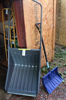 Photo showing both Yard Works snow pusher with wide gray scoop and metal handle alongside blue Suncast plastic snow shovel with handle.