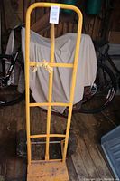 Full view of the yellow steel dolly hand truck standing upright against a wooden backdrop with a covered bicycle behind it.