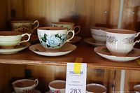 Six assorted vintage teacups and saucers displayed on wood shelf, showing floral patterns and varied colors.