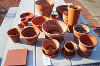 Photo showing an assortment of terracotta pots and saucers arranged on a white picnic table outdoors. Includes various sizes and shapes with natural terracotta color and signs of use.