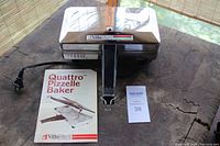 Closed view of stainless steel electric pizzelle baker with black handle and attached cord, placed on wooden surface next to the original recipe and instruction booklet.