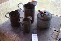 Four vintage tin items on wooden surface: one kerosene jug with wooden handle and three metal measuring tins of different sizes, two marked 'Pint' and 'Quart'.