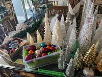 Wide display of assorted Christmas trees of various colors and materials, plastic bins of glass ball ornaments, and garland on a table, clearly showing the variety of decorative trees and ornaments.