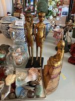 Wide view showing the two gilded Egyptian figurines, golden Buddha figurine, several shells in jars, and decorative stones in front.