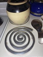 Photo of black and beige ceramic vessel with lid and part of blue kettle next to metal spoon rest on stove coil