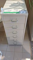 Front view of the six-drawer white metal cabinet showing all drawers and handles, placed on tiled floor.