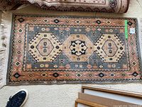 Full view of the handwoven rug placed on carpet showing central medallions and border pattern, off-white fringes visible at one end.