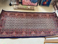 Full view of handwoven runner rug laid out on floor surrounded by other framed artworks and rugs rolled up.
