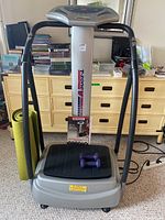 Full front view of the T-Zone VT-12 vibration plate fitness machine showing base platform, vertical support, and digital control panel on top. Purple dumbbells and user's manual placed on the platform.