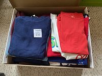 Box filled with folded tops mostly in blue, red, white shades showing tops of garments