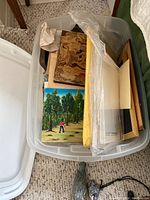 A plastic bin filled with multiple framed artworks, some wrapped, showing a landscape painting with trees and a figure.