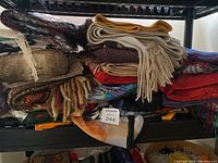 Black metal shelf holding multiple folded scarves stacked in piles. Scarves in various colors including beige, red, brown, mustard yellow, and multicolor patterns.