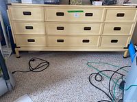 Full view of the yellow dresser showing all nine drawers, black recessed handles, standing on carpeted floor with cords nearby. Shows wear and scratches on surface.
