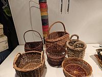 Seven woven baskets displayed together on a surface, showing a variety of shapes and natural color tones along with one colorful tall basket.
