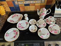The full set of china pieces arranged on a countertop including tea pot, plates, cups, saucers, and other pieces all featuring pink rose floral design with gold trim.