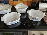 Photo shows 4 white Corning Ware bakeware pieces with blue cornflower pattern on stovetop: 2 with lids and 2 without lids in various shapes including square and rectangular.