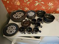 Overall view of the Denby dinnerware set arranged on a white surface with dinner plates, side plates, bowls, cups, and saucers showing the floral pattern and brown glaze cups.