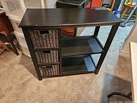 Front and side view of black storage unit with wicker drawers and shelving on carpeted floor.