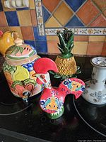 Four ceramic pieces arranged on black surface with colorful tile background: floral jar with lid, pineapple jar, rooster figurine, and white candlestick.