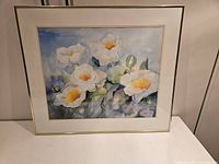 Angled photo of the framed watercolor floral painting showing flowers with white petals and yellow centers, in a light wood frame with cream mat.