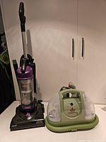 Photo showing side-by-side Bissell AeroSwift upright vacuum and Bissell Little Green steam cleaner on a white surface