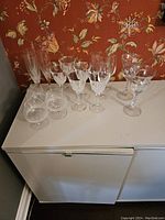 Full collection of 16 crystal stemware pieces arranged on a white cabinet against floral wallpaper background. Various types of glasses including champagne flutes, wine glasses, and cocktail glasses with etched patterns.