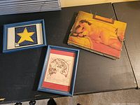 Three small original wall art pieces on black surface including a nude reclining figure on board and two framed pieces with blue frames